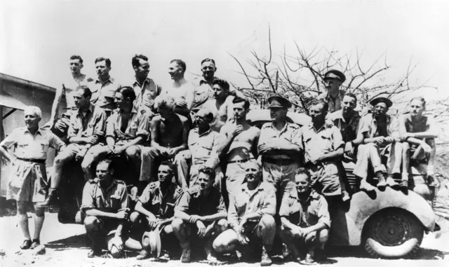 About twenty men in tropical kit posed on and around an open car in hard sun, some standing on it, some sitting on the bonnet, some crouching in front.