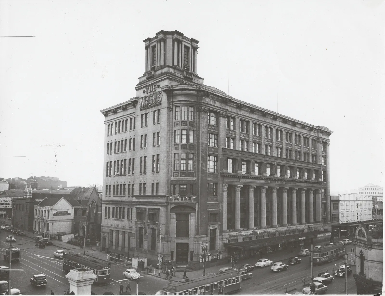 A high view of a pale stone office block on a street corner, seven storeys with a square tower at the corner lettered “The Argus”, a giant colonnade along its long front, trams and cars in the streets below.