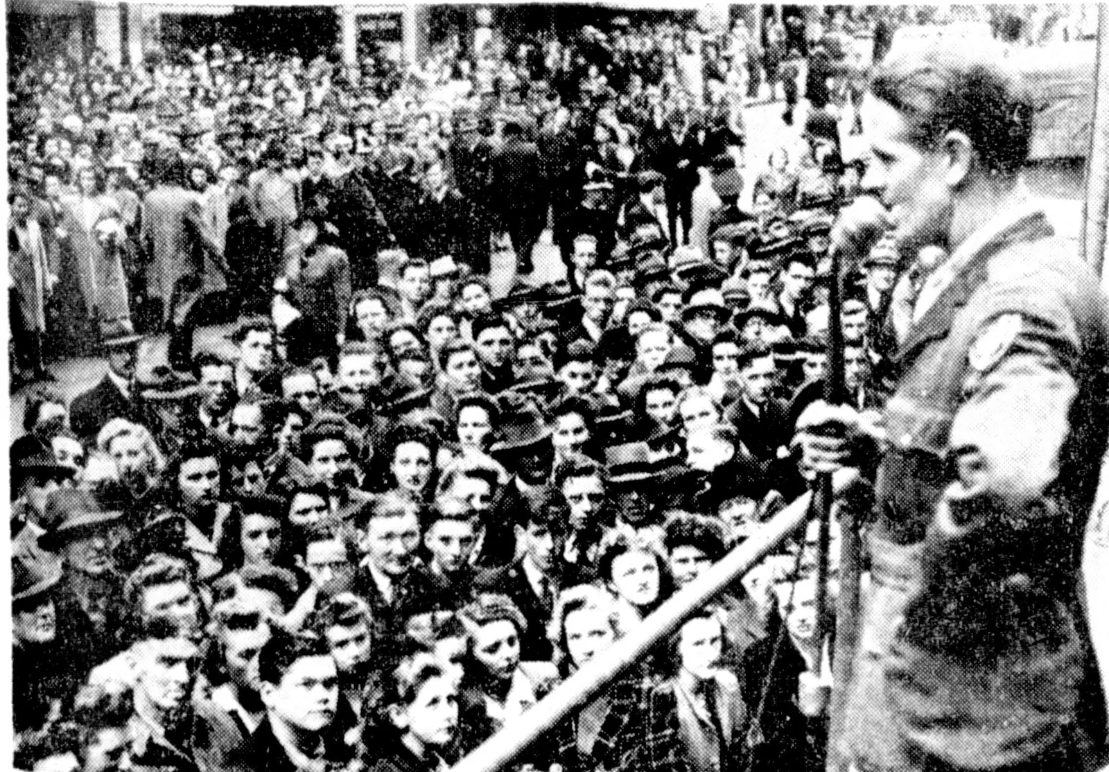 A man in uniform speaks into a microphone from a platform at the right; below him a dense crowd of men and women in hats fills the street, looking up.