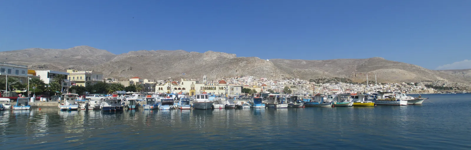 A wide view across a still blue harbour lined with moored fishing boats, the pale houses of a town along the shore and up the slopes, bare grey mountains behind.